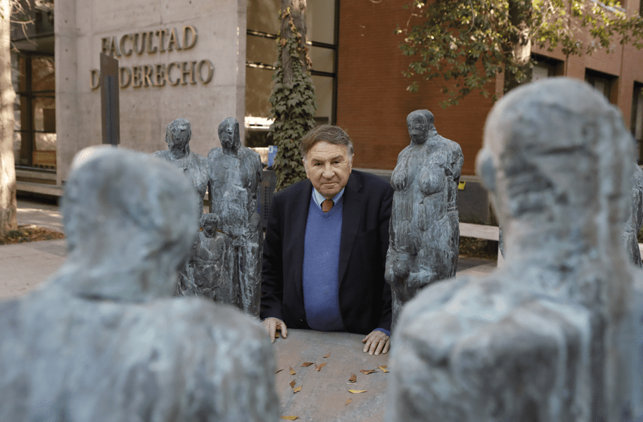 Un hombre mirando de frente, parado detrás de unas estatuas y un edificio al fondo